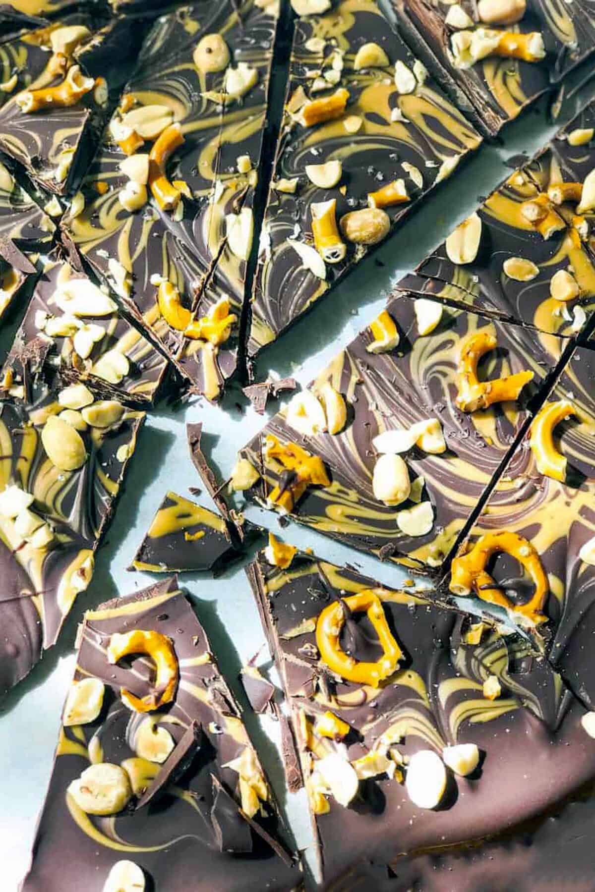 Swirled Chocolate Peanut Butter Bark broken into pieces, topped with cashew nuts, peanuts, and pretzel twists on a white surface.