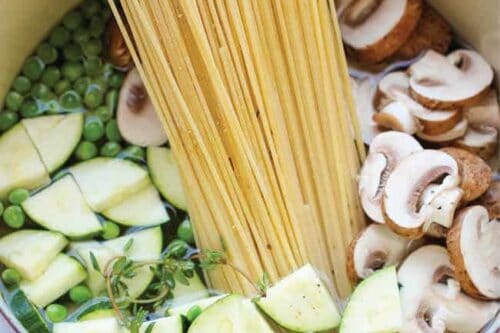 A pot filled with uncooked spaghetti, sliced mushrooms, peas, and chopped zucchini on a marble surface, showcasing a delightful start for zucchini recipes. Fresh thyme is placed on the vegetables, adding a fragrant touch.