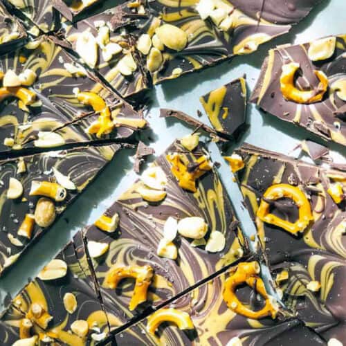 Broken pieces of marbled Chocolate Peanut Butter Bark topped with pretzels, peanuts, and swirls of dark and white chocolate are scattered on a light surface.
