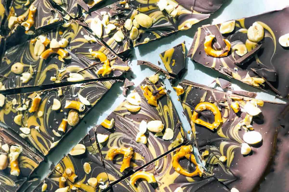 Broken pieces of marbled Chocolate Peanut Butter Bark topped with pretzels, peanuts, and swirls of dark and white chocolate are scattered on a light surface.
