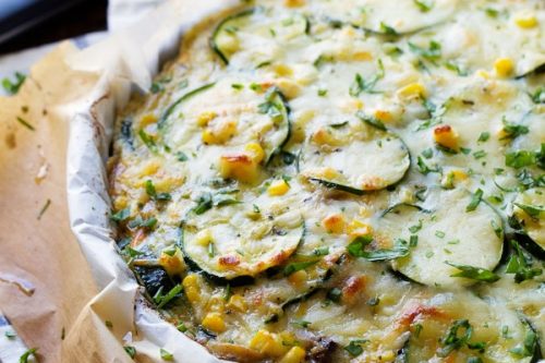 A baked vegetable quiche with sliced zucchini and herbs on top sits on parchment paper, surrounded by a knife and a bowl of chopped herbs-one of the easy zucchini recipes perfect for any meal.