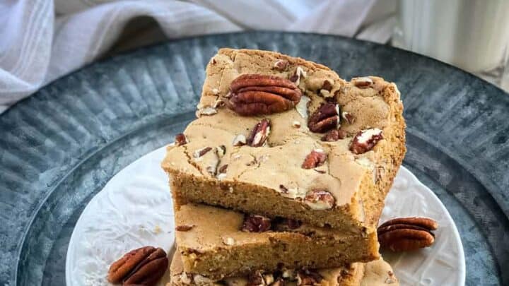 A stack of three maple pecan blondies on a white plate, set on a textured metal charger. The blondies are sprinkled with whole and chopped pecans. A glass of milk and autumn-themed decorations are in the background.