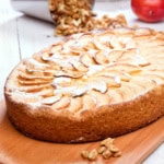 A baked German Apple Kuchen tart sprinkled with powdered sugar sits on a wooden board, with walnuts and an apple in the background.