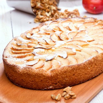 A baked German Apple Kuchen tart sprinkled with powdered sugar sits on a wooden board, with walnuts and an apple in the background.