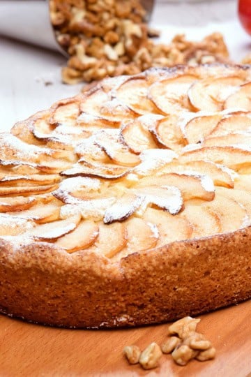 A baked German Apple Kuchen tart sprinkled with powdered sugar sits on a wooden board, with walnuts and an apple in the background.