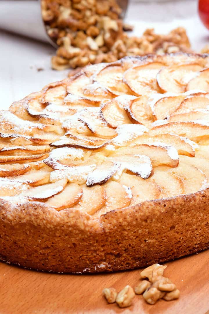 A baked German Apple Kuchen tart sprinkled with powdered sugar sits on a wooden board, with walnuts and an apple in the background.
