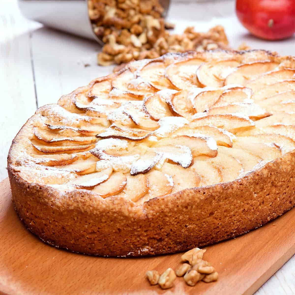 A baked German Apple Kuchen tart sprinkled with powdered sugar sits on a wooden board, with walnuts and an apple in the background.