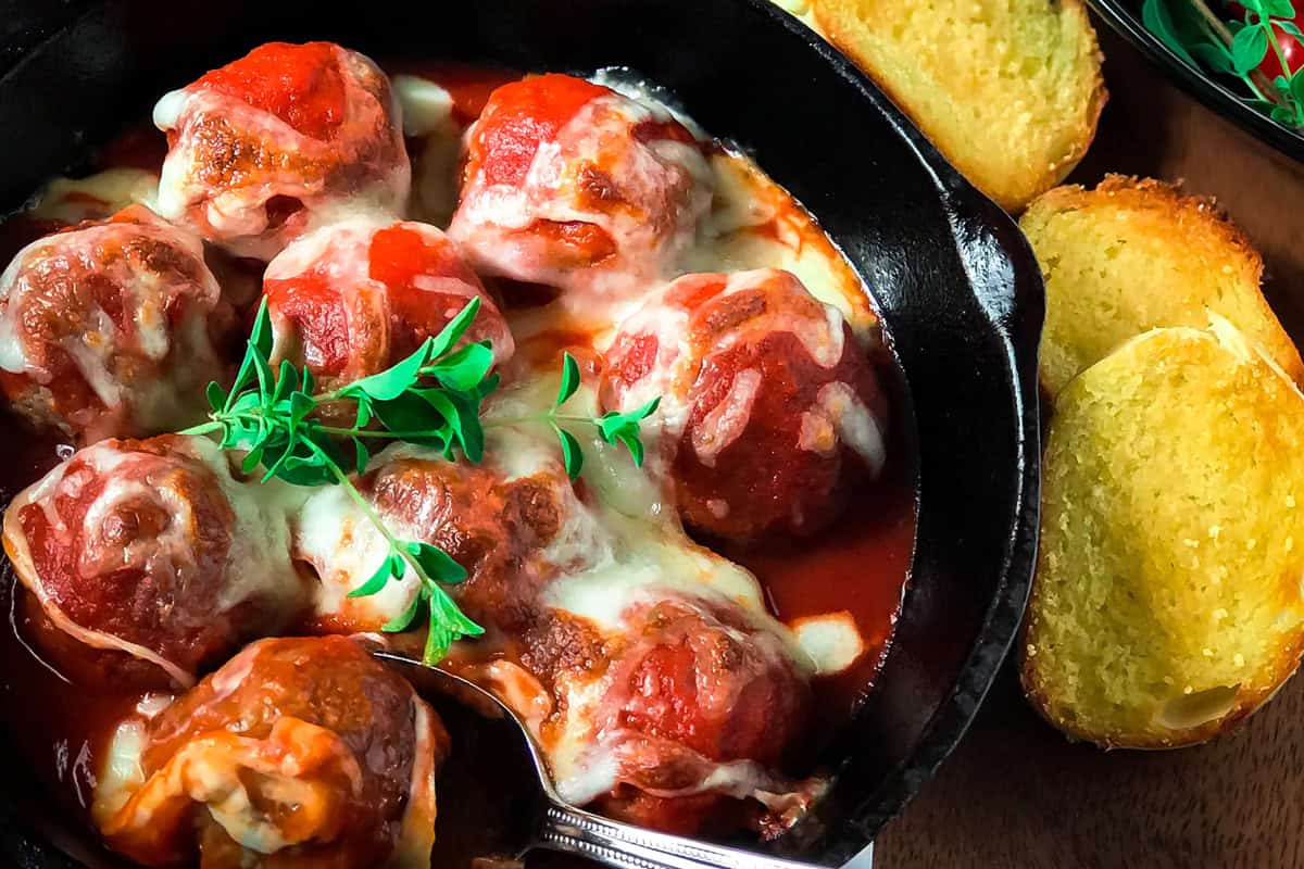 Skillet Meatballs topped with melted cheese and tomato sauce in a black skillet, garnished with fresh herbs, served alongside slices of toasted garlic bread.