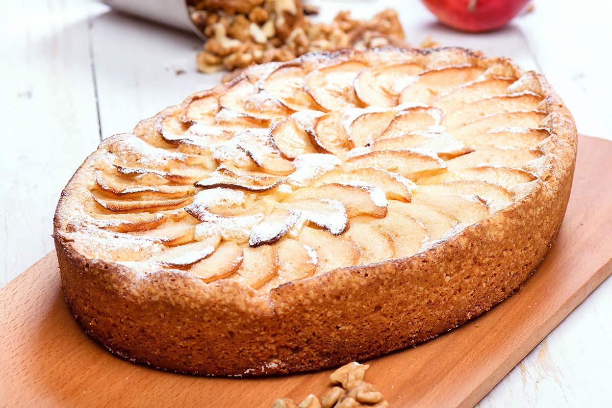 Oval apple tart with thinly sliced apples arranged in a spiral on a wooden board, dusted with powdered sugar; reminiscent of classic German Apple Kuchen. Walnuts scattered nearby.
