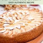 A round German Apple Kuchen topped with thin apple slices and powdered sugar is displayed on a wooden board, with walnuts scattered nearby.