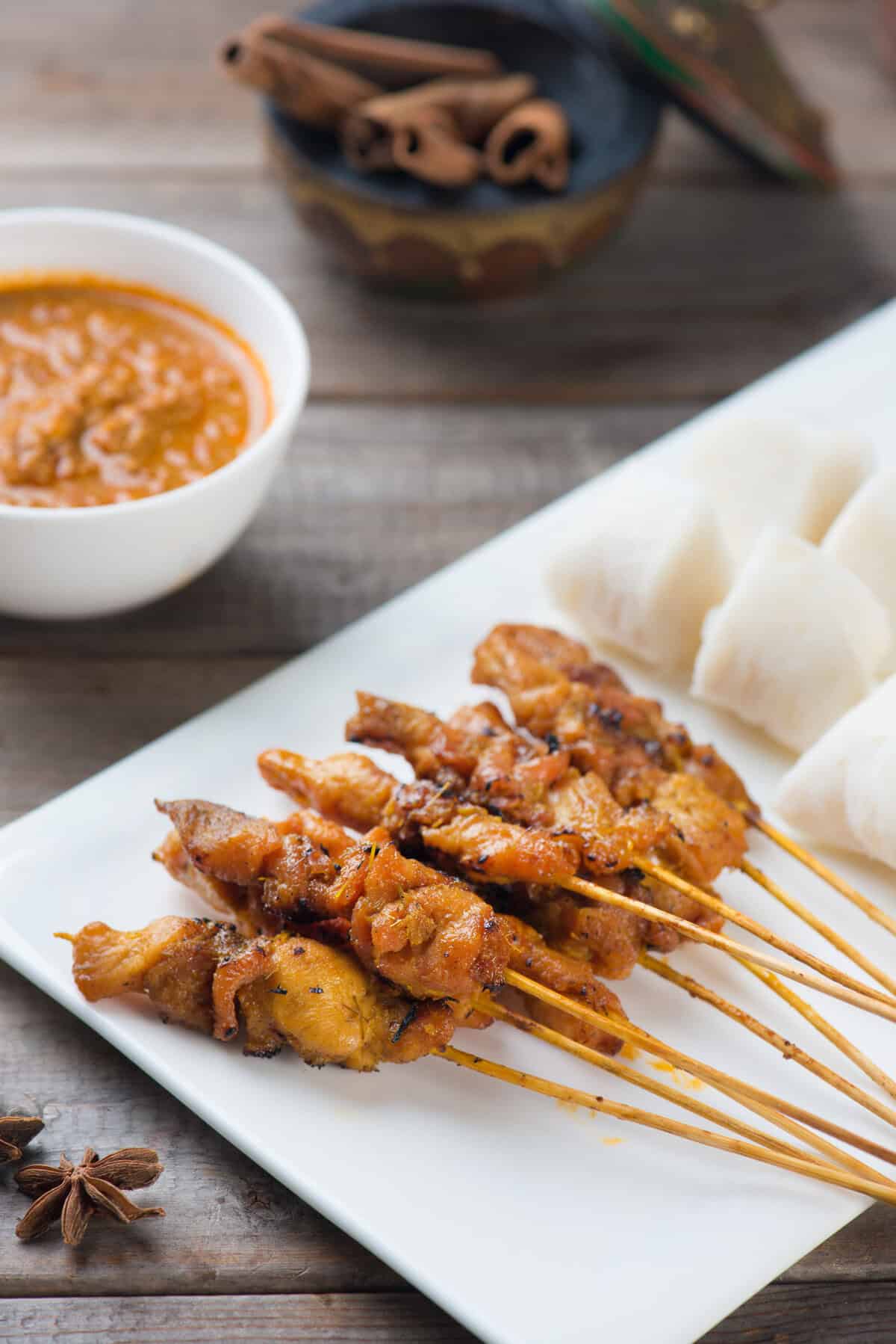 A plate of chicken satay skewers served with cubed rice cakes and a bowl of peanut sauce, placed on a wooden table with aromatic spices in the background.