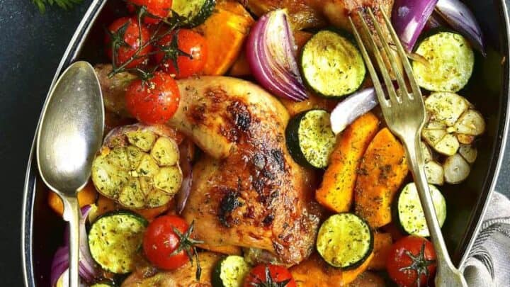 A roasted chicken and vegetable dish in an oval pan features golden-brown chicken hindquarters, cherry tomatoes, sliced zucchini, red onions, and orange bell peppers. A fork and spoon nestle in the pan, with a cloth underneath to complete the inviting scene.