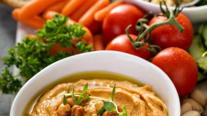 A bowl of red pepper hummus topped with roasted chickpeas and herbs is surrounded by cherry tomatoes, baby carrots, cucumber slices, couscous salad, crackers, parsley, and pistachios on a wooden board.
