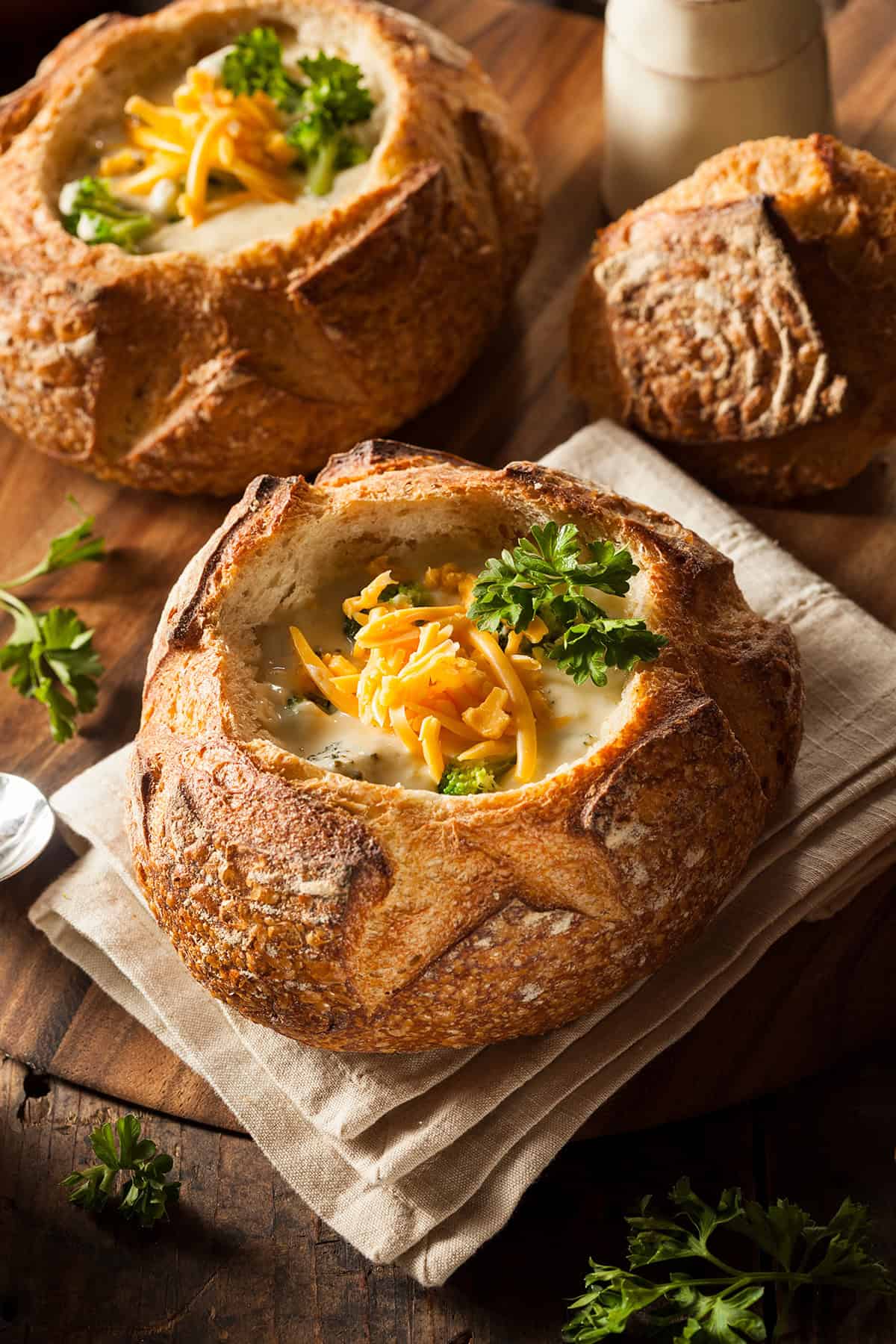 Bread bowl filled with broccoli cheddar soup, garnished with cheddar cheese and curley parsley, with more bowls in the background.