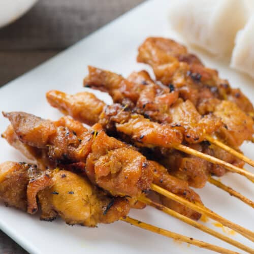 A close-up of chicken satay skewers grilled to perfection and arranged on a white plate, served with pieces of rice cake on the side.