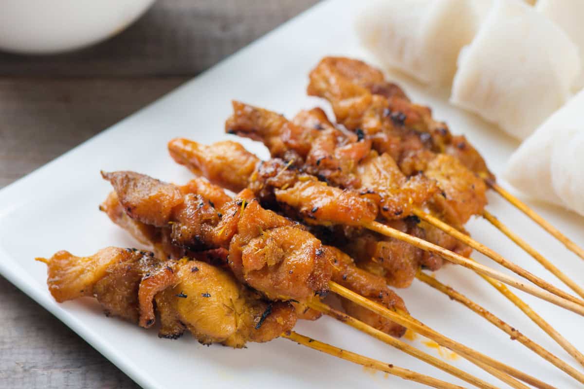 A close-up of chicken satay skewers grilled to perfection and arranged on a white plate, served with pieces of rice cake on the side.