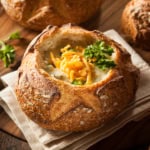 Square image of broccoli cheddar soup in a carved out bread bowl with shredded cheese and curly parsley for garnish.