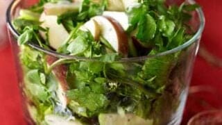 A glass bowl brimming with a Thanksgiving salad, featuring mixed greens and crisp apple slices, is elegantly set against a red tablecloth.