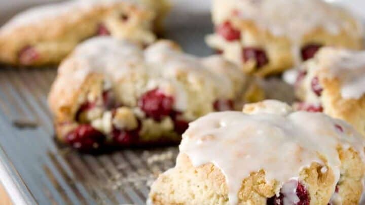 Freshly baked cranberry scones with raspberries and white icing are arranged on a metal baking tray. A spoon with icing rests nearby, and there's a wooden board with baking tools in the blurred background.