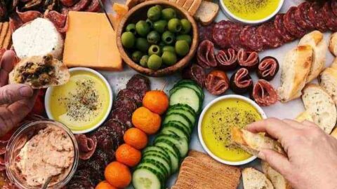 A large Fall Charcuterie Board with assorted crackers, breads, cheese, deli meats, olives, vegetables, nuts, dips, and fruit, with several hands reaching for food.