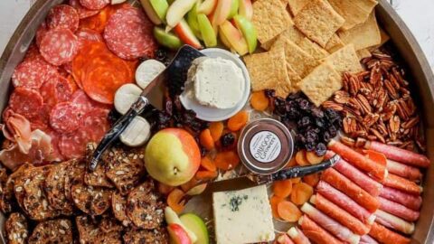 A large round wooden tray with assorted meats, cheeses, crackers, fresh fruit, nuts, and a small jar of jam on a white wooden surface—perfect as a Fall Charcuterie Board for cozy gatherings.