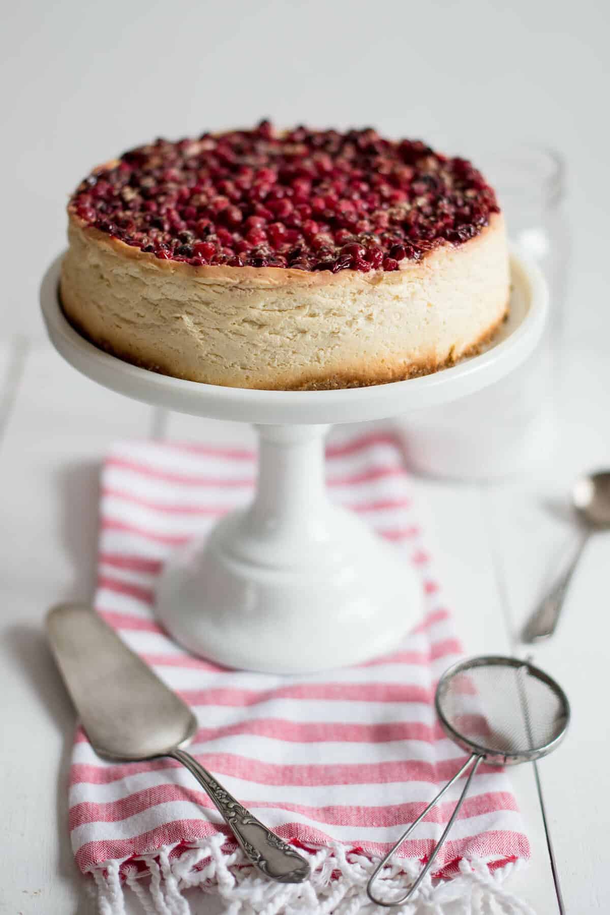 A Cranberry Cheesecake topped with mixed berries sits on a white cake stand, with utensils and a pink striped cloth nearby.