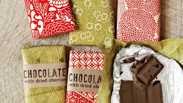 Five wrapped chocolate bars with dried cherries and one unwrapped, partially-eaten bar on a wooden surface—perfect as easy homemade food gifts. The wrappers feature festive red and green patterns.