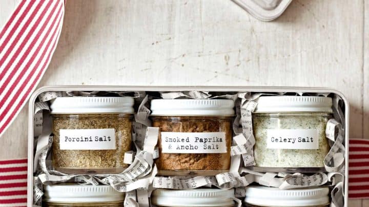 A metal box with six labeled jars of flavored salts—Porcini, Smoked Paprika, Celery, Lime-Ginger, Herbes de Provence, and Curry—displayed with a red-striped ribbon, perfect for easy homemade food gifts.