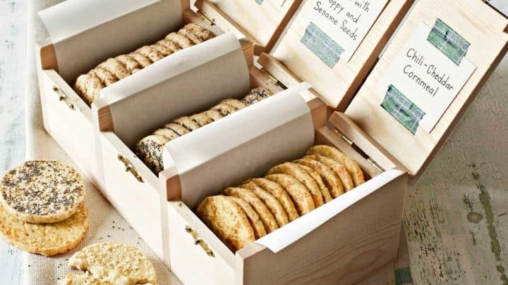 A wooden box with four compartments displays assorted crackers, each labeled with a flavor card. Some crackers are placed on a striped cloth beside the box—perfect as easy homemade food gifts.