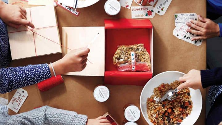 Four people are packaging easy homemade food gifts—granola into boxes, adding handwritten holiday gift tags, tying red string, and using white jars on a brown paper-covered table.
