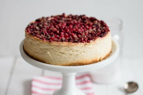 Horizontal view of unsliced Cranberry Cheesecake on a white cake stand resting on a red and white striped kitchen linen in front of a white neutral background.