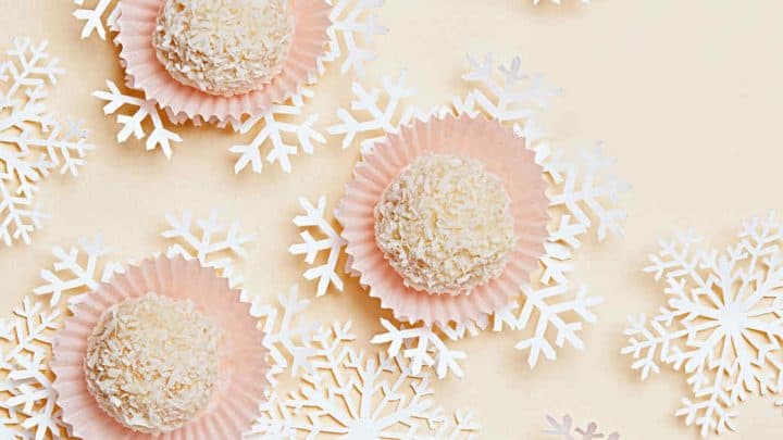 Five round coconut-covered candies in pink paper cups are arranged on a light background with decorative white paper snowflakes—perfect for easy homemade food gifts.