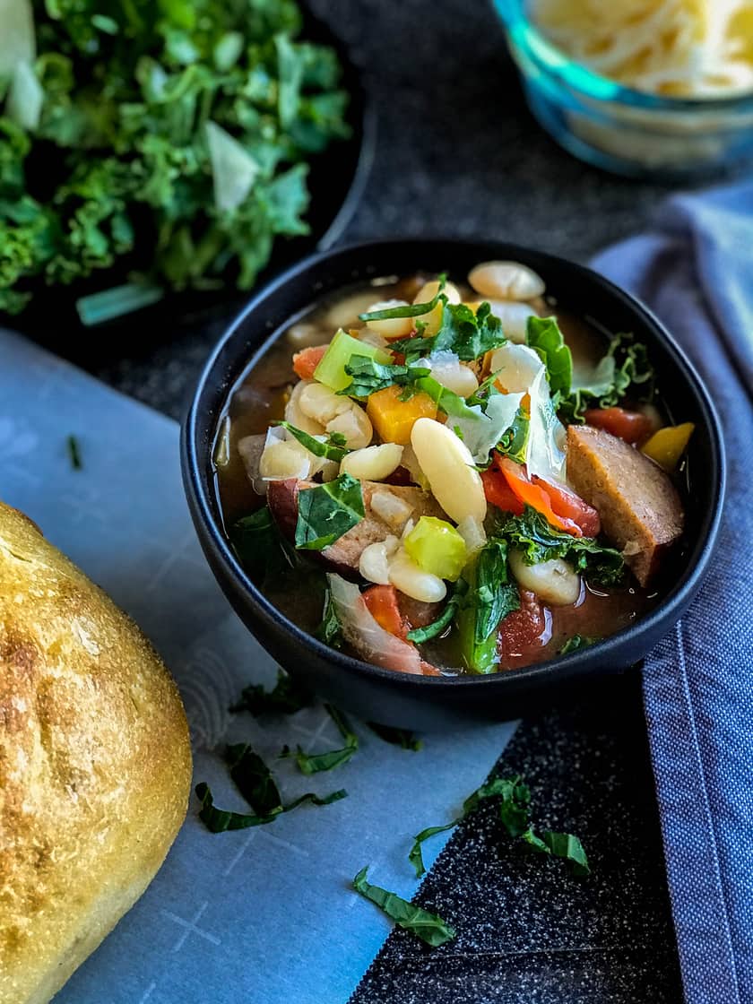 Top view of White Bean Kielbasa and Kale Soup served with a fresh baguette in a black bowl with fresh herbs on the side.