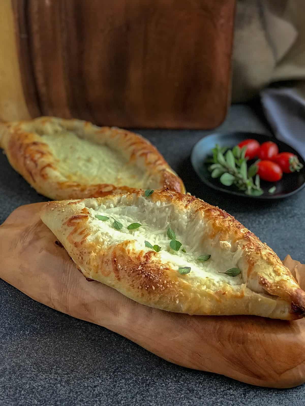 Two baked Turkish cheese boat pide breads with melted cheese filling rest on a wooden board, with a plate of cherry tomatoes and herbs in the background.