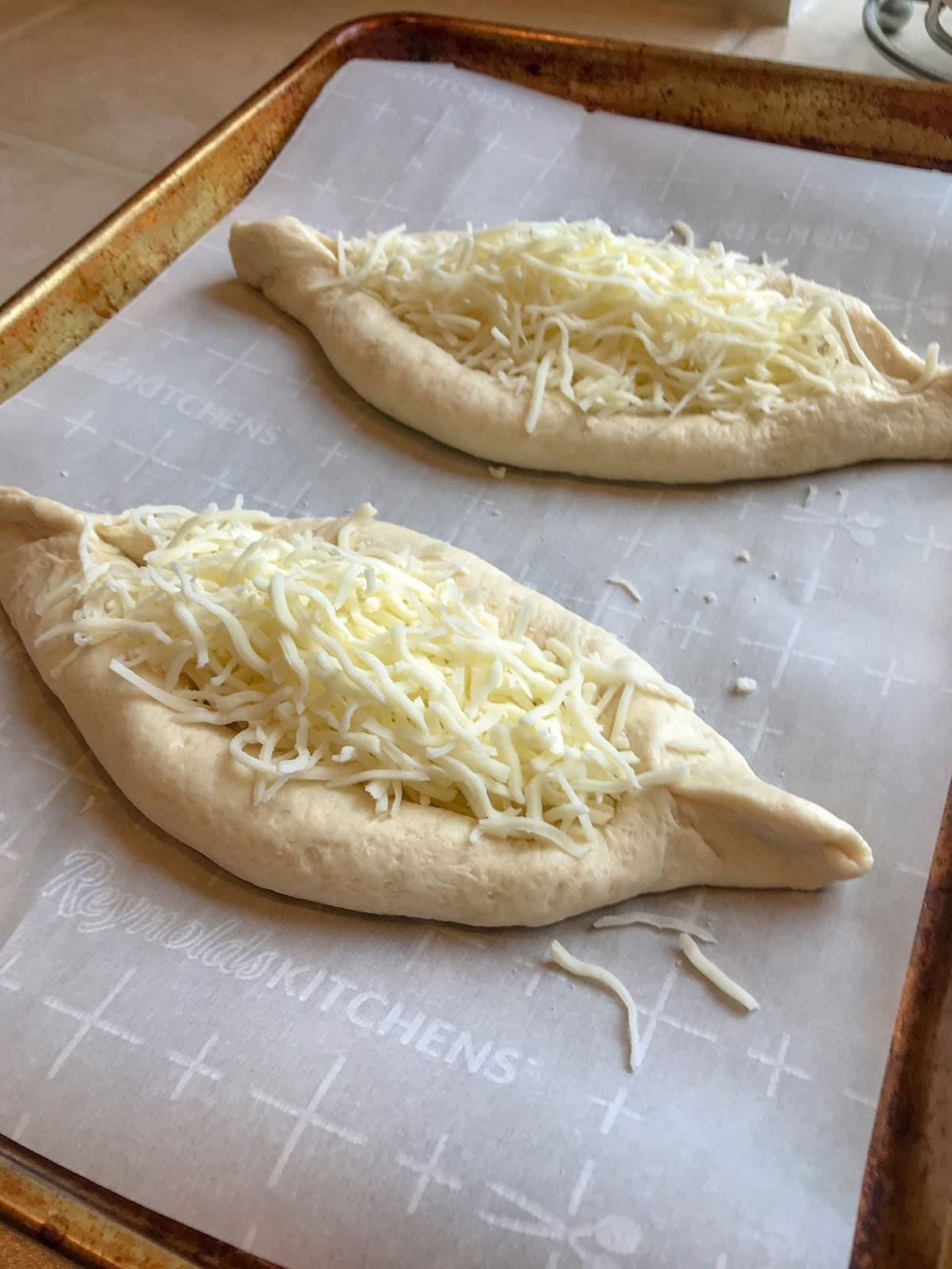 Two uncooked cheese boats, shaped like small boats and topped with shredded cheese, are placed on a parchment-lined baking sheet.