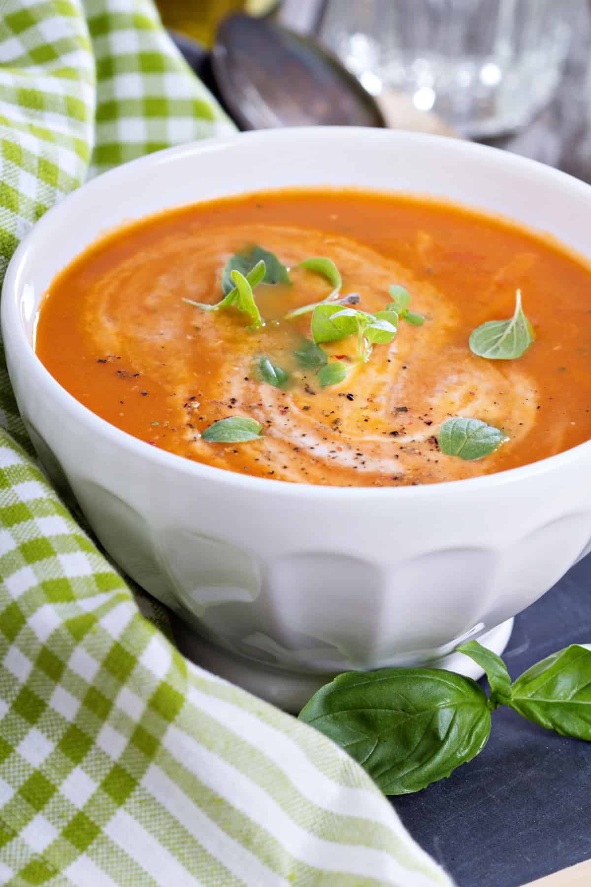 Bowls of garden tomato soup garnish with small microgreens, and black pepper rests in a white bowl with a checkered cloth nearby.