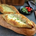 Two baked cheese boat Turkish pide breads rest on a wooden board, with a small plate of cherry tomatoes and herbs in the background.