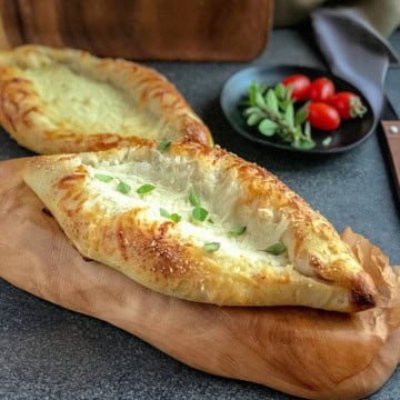 Two baked cheese boat Turkish pide breads rest on a wooden board, with a small plate of cherry tomatoes and herbs in the background.