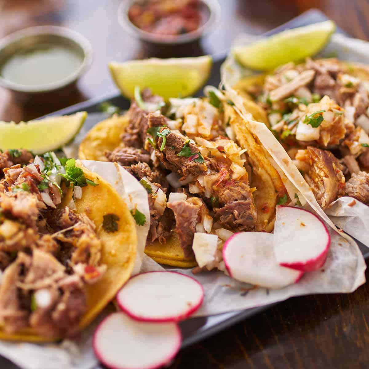 Three tacos filled with Mexican shredded chicken, topped with onions and cilantro, served with lime wedges, sliced radishes.