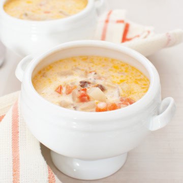 A white soup bowl filled with creamy salmon chowder, featuring visible pieces of potato, carrot, and possibly meat, sits on a matching white surface with a striped napkin nearby.