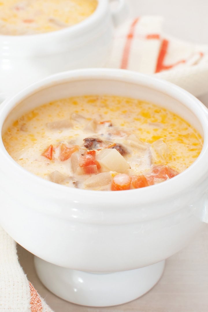 A white soup bowl filled with creamy salmon chowder, featuring visible pieces of potato, carrot, and possibly meat, sits on a matching white surface with a striped napkin nearby.