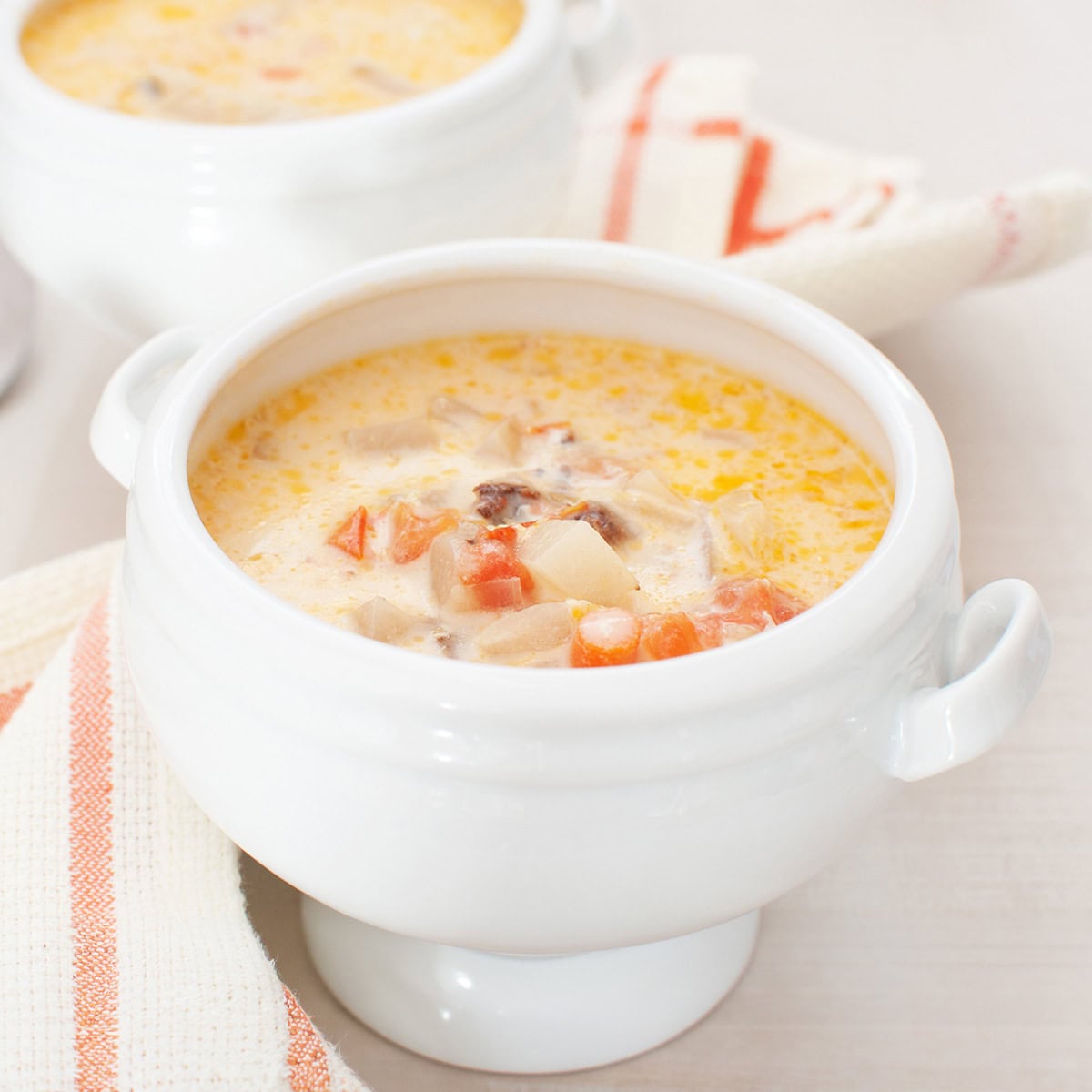 A white soup bowl filled with creamy salmon chowder, featuring visible pieces of potato, carrot, and possibly meat, sits on a matching white surface with a striped napkin nearby.