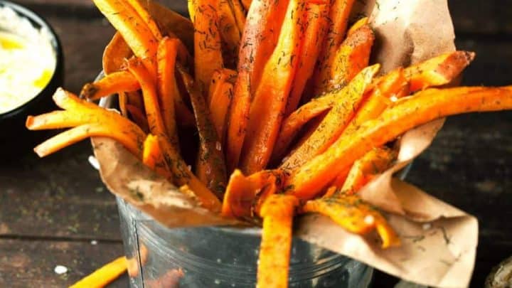 A metal container cradles piping hot sweet potato fries, impeccably crispy and seasoned with fragrant herbs, lined with brown paper. Nearby, garlic cloves and salt crystals are artfully scattered on a rustic wooden surface.