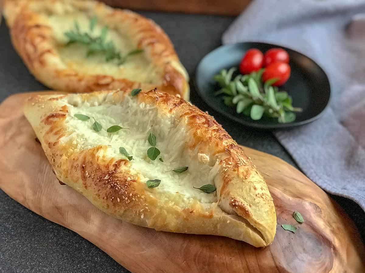 Two baked Turkish cheese boat pide breads with melted cheese and herbs rest on a wooden board, with a small plate of tomatoes and greens in the background.