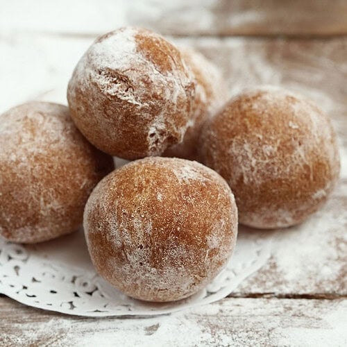 Five round, powdered sugar-covered baked doughnut holes are stacked on a white doily atop a wooden surface lightly dusted with sugar.