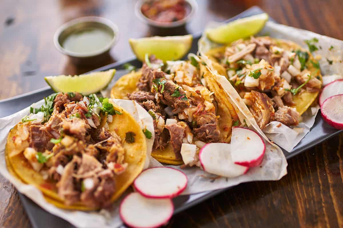 Three Mexican shredded chicken tacos topped with chopped onion and cilantro are served with lime wedges, sliced radishes, and sauces on a rectangular tray. 