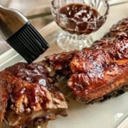 On a white tray, a barbecued rack of slow cooker ribs is partially sliced, while a basting brush applies sauce. In the background, a small glass dish brims with rich barbecue sauce.