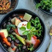 A bowl of colorful salad with black beans, greens, sliced vegetables, and a dollop of cream garnished with herbs evokes the hearty essence of black bean soup. Nearby are a bowl of red pepper flakes, a napkin, and two spoons on a light surface.