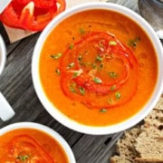 Two bowls of red pepper soup garnished with red bell pepper slices and herbs. A spoon is beside one bowl, and slices of bread are nearby on a wooden table.
