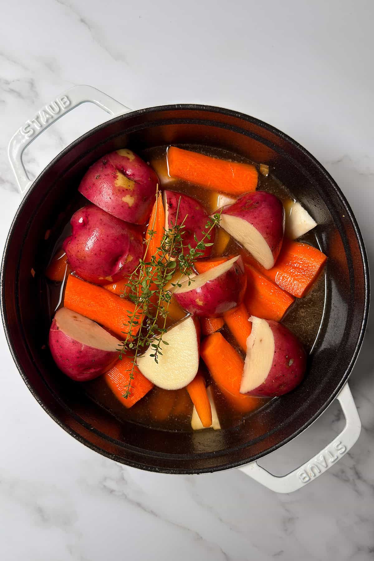 A pot containing halved red potatoes, chopped carrots, garlic cloves, fresh thyme, and broth on a white marble surface-classic ingredients for a hearty Irish Stew.